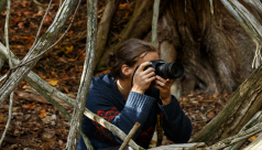 A woman crouches in the woods, her camera raised to her face, ready to take a photo.